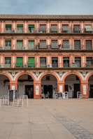 A multi-story building with a repeating pattern of windows and green shutters, accompanied by a series of archways at the ground level. The structure features a Mediterranean architectural style with a warm, earthy color palette. A few people are seated at outdoor tables near the arches, and stacks of chairs are arranged on the paved courtyard in front.
