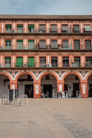 A multi-story building with a repeating pattern of windows and green shutters, accompanied by a series of archways at the ground level. The structure features a Mediterranean architectural style with a warm, earthy color palette. A few people are seated at outdoor tables near the arches, and stacks of chairs are arranged on the paved courtyard in front.