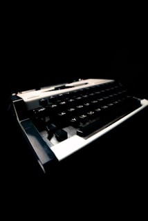 A close-up view of a vintage typewriter featuring a slightly angled perspective. The focus is on the keys, which are highlighted against a dark background, creating a stark contrast. The typewriter is partially illuminated, accentuating its classic design.