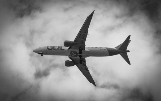 An airplane captured from below, flying against a backdrop of clouds. The image is in black and white, enhancing the contrast between the aircraft and the cloudy sky.