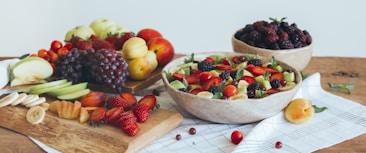 An assortment of fresh fruits is displayed on a wooden table. There are sliced and whole apples, bananas, strawberries, grapes, blackberries, and apricots. The colorful arrangement is spread over two wooden cutting boards and a bowl, with some fruits placed directly on a white checkered cloth.