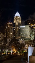 A nighttime cityscape featuring several illuminated skyscrapers. The tallest building is prominently lit with bright white and blue lights. A sign with 'wework' is visible on one of the buildings. Trees with decorative lights line a walkway in the foreground, adding a warm contrast to the cool tones of the buildings.