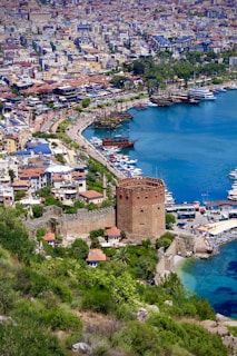 The image depicts a coastal city with a prominent historic tower and fortress at the forefront. A marina with various boats and ships is visible along the vibrant turquoise waters, contrasting with the lush greenery surrounding the fortification. The backdrop showcases a densely populated urban area with numerous buildings spread across the landscape.