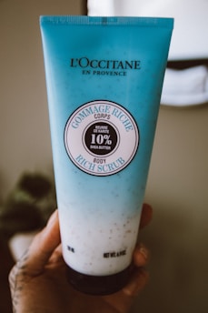 A blue tube of L'Occitane body scrub is held in a hand. The tube features the label 'Gommage Riche Corps' with 10% shea butter indicated. The background is softly blurred, highlighting the product.