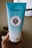 A blue tube of L'Occitane body scrub is held in a hand. The tube features the label 'Gommage Riche Corps' with 10% shea butter indicated. The background is softly blurred, highlighting the product.