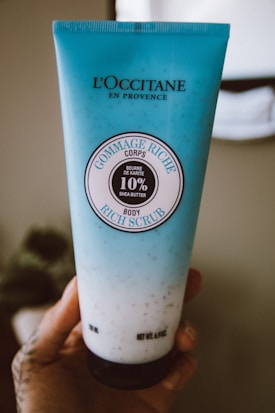 A blue tube of L'Occitane body scrub is held in a hand. The tube features the label 'Gommage Riche Corps' with 10% shea butter indicated. The background is softly blurred, highlighting the product.