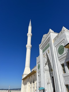 A grand mosque with a tall, white minaret against a clear blue sky. The mosque features intricate architectural designs, including ornate carvings and green circular decorations near the entrance. People can be seen walking near the base of the structure, some of whom are shaded by a small umbrella.