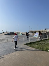A skate park with several people engaging in activities like skating and walking. The area is open with a clear blue sky overhead. A few ramps and rails are visible, covered in graffiti. The atmosphere seems lively and sunny.