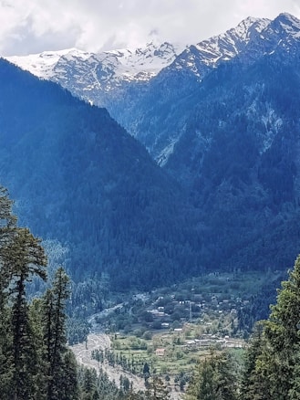 A serene landscape featuring snow-capped mountains in the background, dense green forests covering the slopes, and a small village nestled in a lush valley. The sky is partly cloudy, adding depth to the scene.