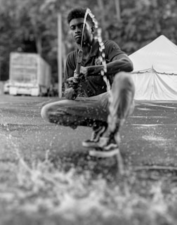A person is crouched down, holding a hose with water spraying out onto the ground, producing splashes. The background includes a tent and a truck, with a natural setting behind.
