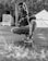 A person is crouched down, holding a hose with water spraying out onto the ground, producing splashes. The background includes a tent and a truck, with a natural setting behind.