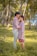 A couple stands closely together in a lush outdoor setting with many palm trees. The man gently kisses the woman's forehead while she holds a bouquet of pink and white flowers. Both are dressed in light shades, and the sunlight filters softly through the trees, creating a serene atmosphere.