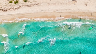 Aerial view of a sandy beach with clear turquoise waters. Several people are visible, some engaging in water activities like kite surfing. Waves gently crash onto the shore, and kites with colorful sails can be seen above the water. A few beachgoers are scattered along the sand, enjoying the sunny day.