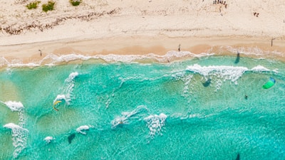 Aerial view of a sandy beach with clear turquoise waters. Several people are visible, some engaging in water activities like kite surfing. Waves gently crash onto the shore, and kites with colorful sails can be seen above the water. A few beachgoers are scattered along the sand, enjoying the sunny day.