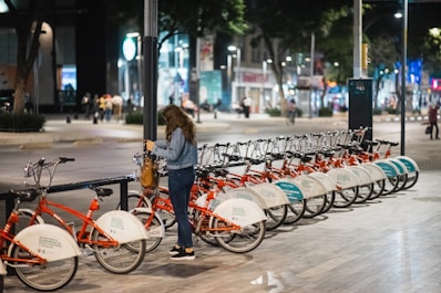 A woman stands near a row of parked red rental bicycles on a nighttime city street, with other people and buildings visible in the background. The scene is illuminated by streetlights and city lights.