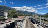 A train station platform is pictured, bordered by railway tracks extending into the distance. Residential buildings are visible on the left side, while a row of trees and a few red buses add color to the scene. Behind the station, a mountain range rises majestically under a sky dotted with scattered white clouds.