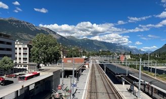 A train station platform is pictured, bordered by railway tracks extending into the distance. Residential buildings are visible on the left side, while a row of trees and a few red buses add color to the scene. Behind the station, a mountain range rises majestically under a sky dotted with scattered white clouds.