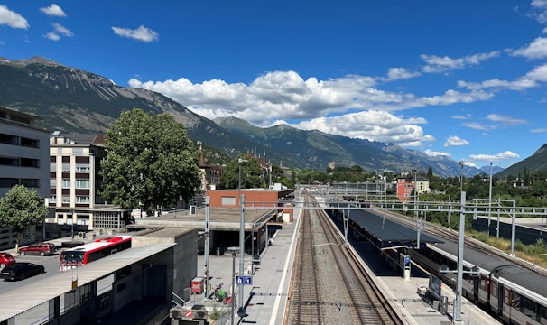 A train station platform is pictured, bordered by railway tracks extending into the distance. Residential buildings are visible on the left side, while a row of trees and a few red buses add color to the scene. Behind the station, a mountain range rises majestically under a sky dotted with scattered white clouds.