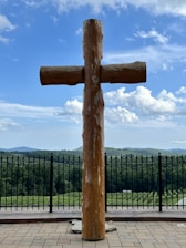 A large wooden cross stands prominently on a stone platform, with a background of lush green hills and a blue, cloud-dotted sky. Black wrought iron fencing is visible behind the cross, providing a boundary at the edge of the platform.