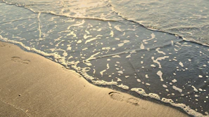 Waves gently crashing on the shore with footprints leading to a sunlit boardwalk and a hint of a fashion magazine peeking out