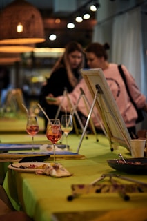Two people, possibly participating in a painting workshop, are positioned near easels on a table that is set up for an art activity. The table is adorned with glasses containing a pinkish drink, paintbrushes, and art materials. Warm lighting adds a cozy ambiance to the setting.