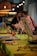 Two people, possibly participating in a painting workshop, are positioned near easels on a table that is set up for an art activity. The table is adorned with glasses containing a pinkish drink, paintbrushes, and art materials. Warm lighting adds a cozy ambiance to the setting.