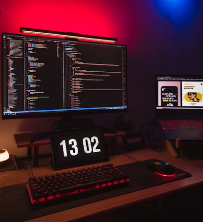 A computer setup featuring a large monitor displaying lines of colorful code in a dark theme. Beneath the monitor, a digital clock reads 13:02. A mechanical keyboard and a gaming mouse rest on a mouse pad illuminated by red backlighting. To the side, a laptop displays web content with a bright, well-lit interface. The room is dimly lit, with ambient red and blue LED lights.