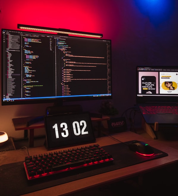 A computer setup featuring a large monitor displaying lines of colorful code in a dark theme. Beneath the monitor, a digital clock reads 13:02. A mechanical keyboard and a gaming mouse rest on a mouse pad illuminated by red backlighting. To the side, a laptop displays web content with a bright, well-lit interface. The room is dimly lit, with ambient red and blue LED lights.