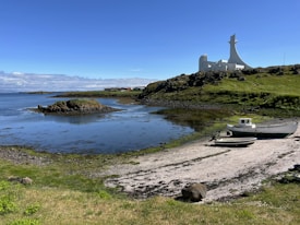 A serene coastal landscape featuring a modern white structure resembling a church on a grassy hill overlooking the water. Two boats are positioned on a small pebbled beach in the foreground. The sky is predominantly clear with some clouds on the horizon.