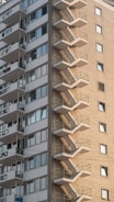 A tall residential building with a prominent external staircase running along the side. The facade features a mix of brick and concrete with numerous windows, some with balconies.