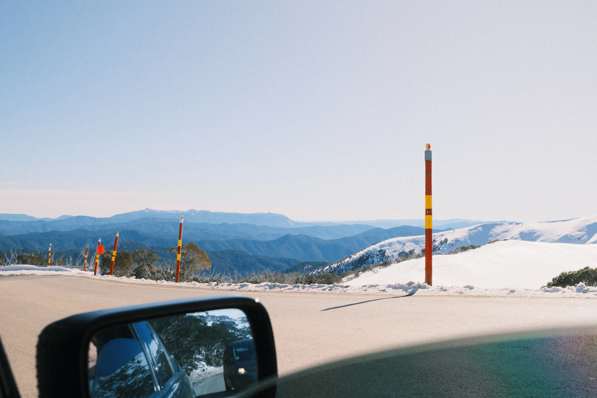 A snowy mountain landscape viewed from a roadside, with snow poles, a winding road and a car side mirror in the foreground.