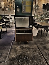 A vintage television set is prominently displayed in a dimly lit industrial space with exposed brick walls. The area features a mix of modern and rustic elements, including various tables and chairs, plants, and wall decor that includes informational posters and a QR code. The floor appears worn, adding to the retro and nostalgic atmosphere.