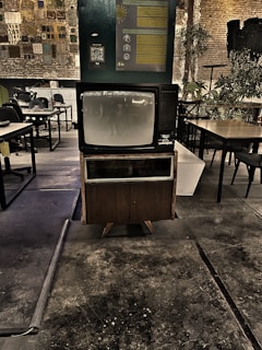 A vintage television set is prominently displayed in a dimly lit industrial space with exposed brick walls. The area features a mix of modern and rustic elements, including various tables and chairs, plants, and wall decor that includes informational posters and a QR code. The floor appears worn, adding to the retro and nostalgic atmosphere.