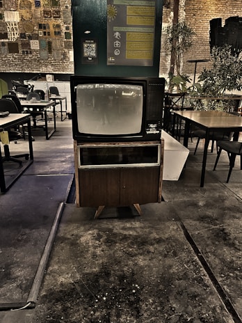 A vintage television set is prominently displayed in a dimly lit industrial space with exposed brick walls. The area features a mix of modern and rustic elements, including various tables and chairs, plants, and wall decor that includes informational posters and a QR code. The floor appears worn, adding to the retro and nostalgic atmosphere.