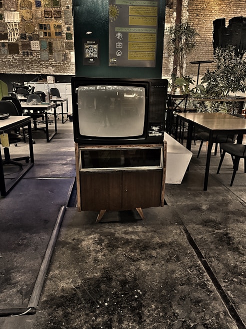 A vintage television set is prominently displayed in a dimly lit industrial space with exposed brick walls. The area features a mix of modern and rustic elements, including various tables and chairs, plants, and wall decor that includes informational posters and a QR code. The floor appears worn, adding to the retro and nostalgic atmosphere.