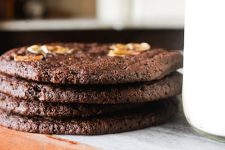 A stack of thick, homemade chocolate cookies with visible texture and embedded nuts or chocolate chips. The cookies are placed on a wooden surface next to a glass jar filled with milk.