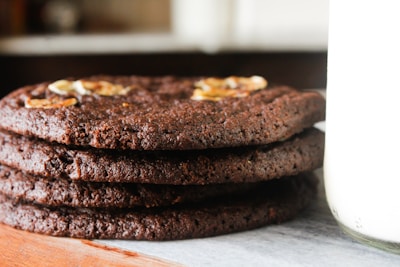 A stack of thick, homemade chocolate cookies with visible texture and embedded nuts or chocolate chips. The cookies are placed on a wooden surface next to a glass jar filled with milk.