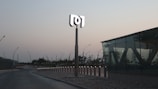 A modern, minimalistic outdoor area features a tall sign with a stylized logo on top, surrounded by sleek street lamps lining a curved roadway. The setting is dusk, with a backdrop of trees and buildings under a clear sky. A glass structure with angular designs is prominently displayed on the right, adding a contemporary architectural element.