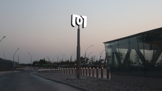 A modern, minimalistic outdoor area features a tall sign with a stylized logo on top, surrounded by sleek street lamps lining a curved roadway. The setting is dusk, with a backdrop of trees and buildings under a clear sky. A glass structure with angular designs is prominently displayed on the right, adding a contemporary architectural element.
