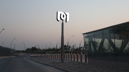 A modern, minimalistic outdoor area features a tall sign with a stylized logo on top, surrounded by sleek street lamps lining a curved roadway. The setting is dusk, with a backdrop of trees and buildings under a clear sky. A glass structure with angular designs is prominently displayed on the right, adding a contemporary architectural element.