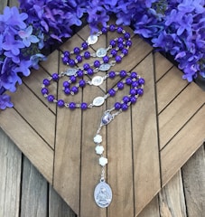 Purple rosary beads are placed on a wooden surface, surrounded by vibrant purple flowers. The rosary features silver medallions and a larger medallion at the end with religious imagery.