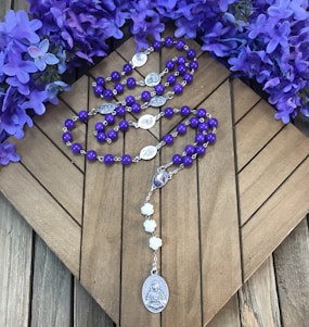 Purple rosary beads are placed on a wooden surface, surrounded by vibrant purple flowers. The rosary features silver medallions and a larger medallion at the end with religious imagery.