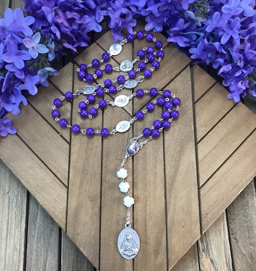 Purple rosary beads are placed on a wooden surface, surrounded by vibrant purple flowers. The rosary features silver medallions and a larger medallion at the end with religious imagery.