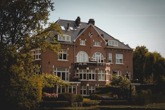 A large, elegant brick house with multiple windows and a dark, tiled roof surrounded by trees and lush greenery. The architecture features arched and rectangular windows, with a symmetrical facade and a small balcony adorned with potted flowers. The house is situated in a serene setting, with a small garden in the foreground.