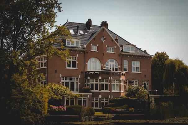 A large, elegant brick house with multiple windows and a dark, tiled roof surrounded by trees and lush greenery. The architecture features arched and rectangular windows, with a symmetrical facade and a small balcony adorned with potted flowers. The house is situated in a serene setting, with a small garden in the foreground.