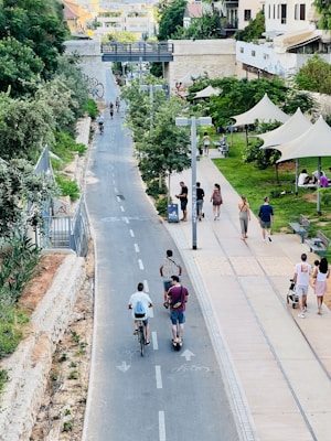 A vibrant urban scene features a paved path lined with trees and greenery. People are strolling, riding bicycles, and using scooters. Benches and shaded areas are visible on the right, where groups relax and socialize. In the background, a bridge and buildings enhance the cityscape.