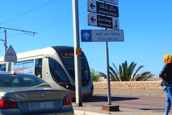A modern tram is passing through an urban area with clear blue skies. There are several traffic signs in multiple languages pointing to local attractions. A woman wearing a bright yellow headscarf and dark jacket stands on the sidewalk. A car is also visible on the road with a license plate displayed.