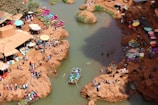 An overhead view of a lively riverside location, with numerous people gathered on rocky terrain alongside the water. Several colorful stalls and umbrellas line the area, adding vibrancy to the scene. A boat with passengers is visible on the river, and a few individuals are swimming. The surroundings exhibit a bustling, festive atmosphere.