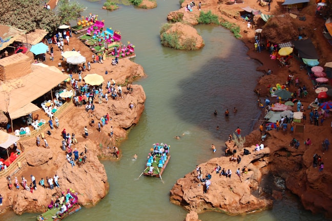 An overhead view of a lively riverside location, with numerous people gathered on rocky terrain alongside the water. Several colorful stalls and umbrellas line the area, adding vibrancy to the scene. A boat with passengers is visible on the river, and a few individuals are swimming. The surroundings exhibit a bustling, festive atmosphere.