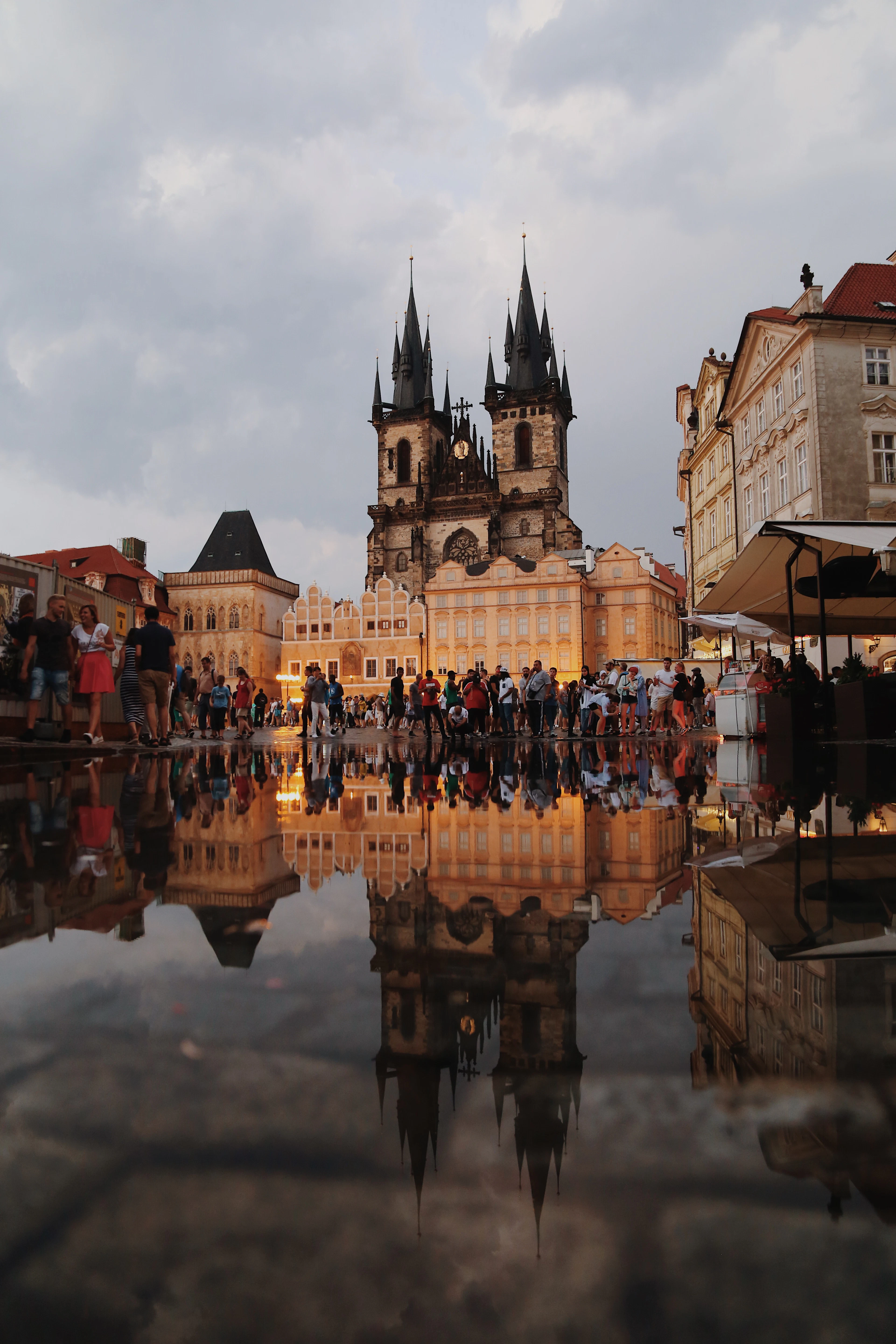 Tyn Church glows gold at dusk over Old Town Square, its towers reflected in a rain puddle among the crowd.